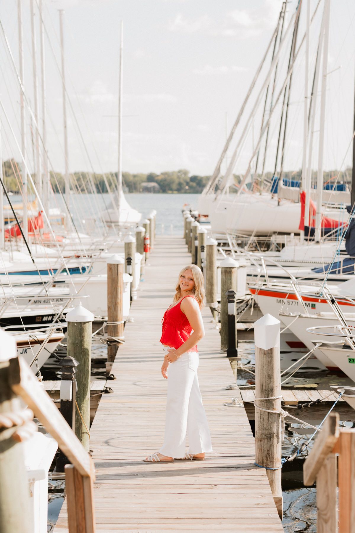 Blonde woman in red top and white pants standing on sunny marina dock with sailboats behind her.