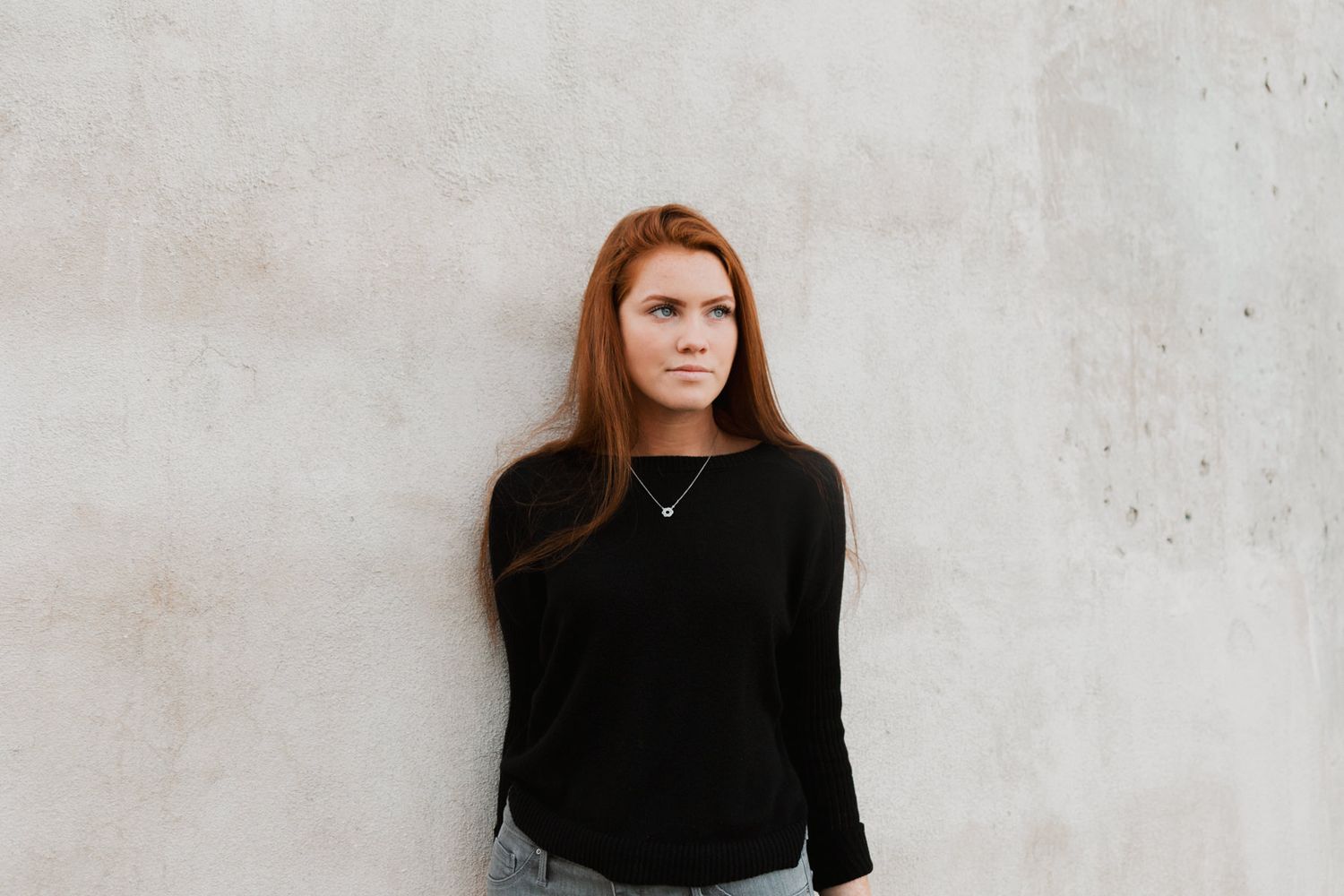 Young woman with long red hair wearing a black long-sleeve top, posing against a light concrete wall.