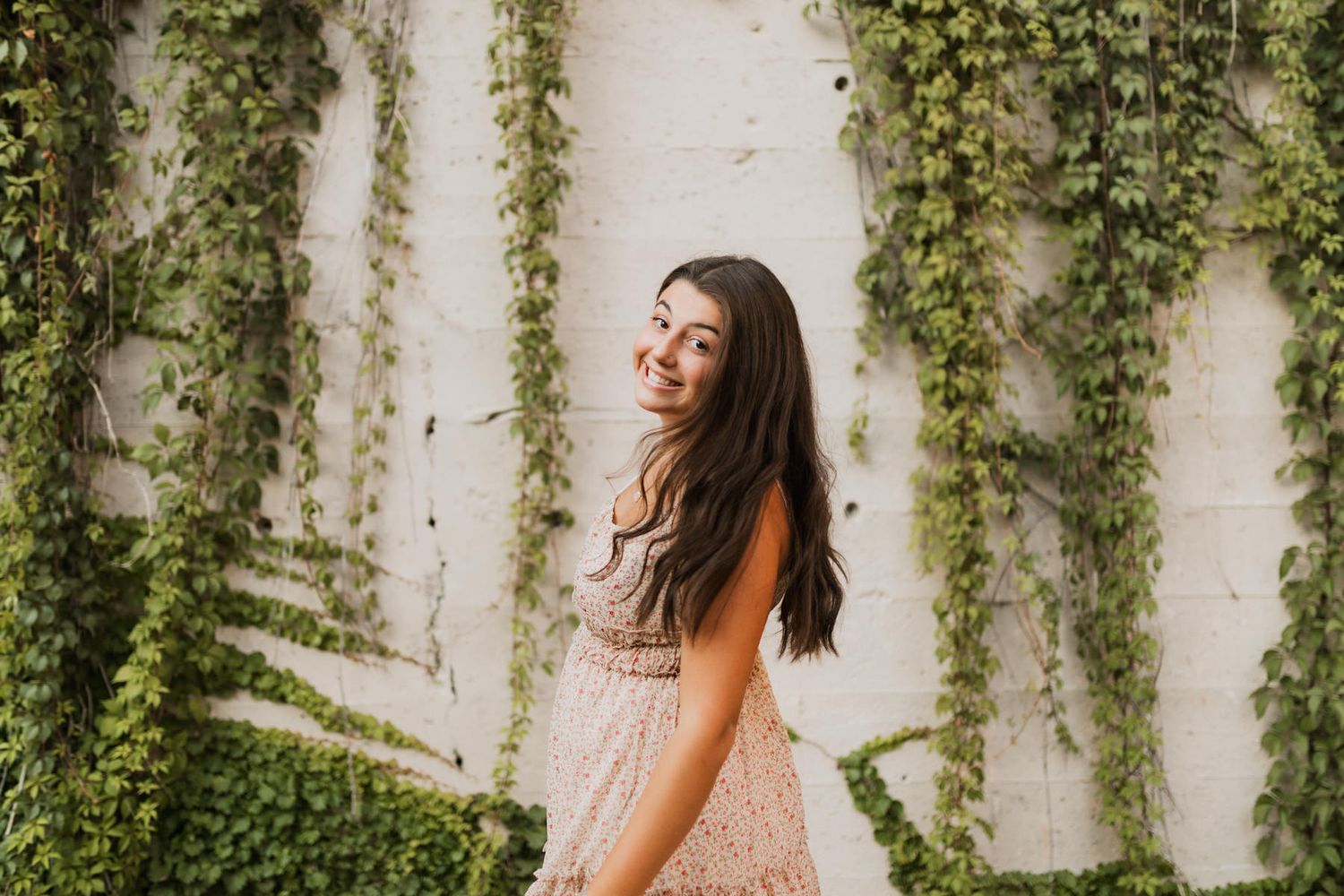 Smiling brunette woman in pink lace dress poses against white wall covered in green ivy.