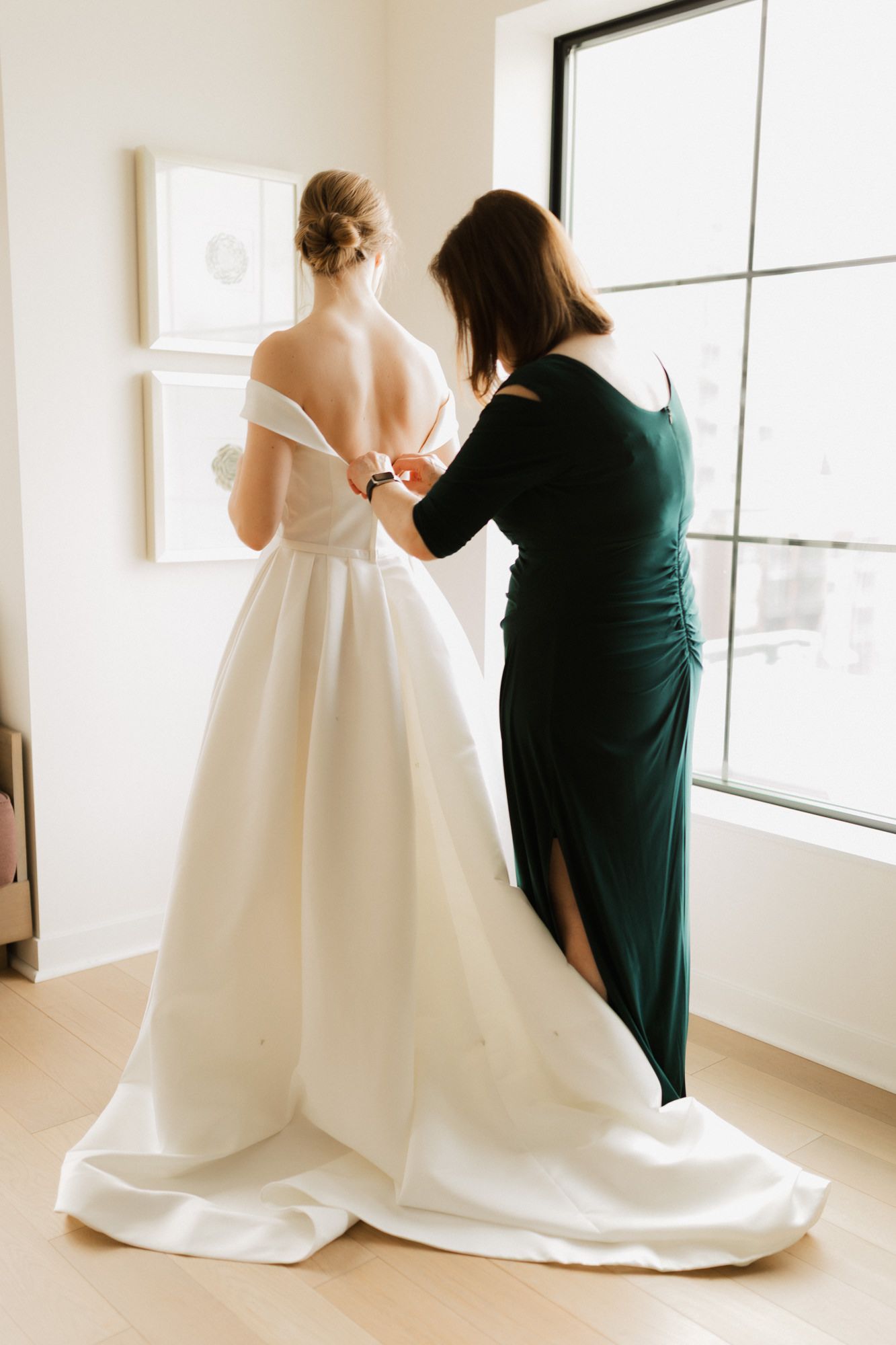 A woman in a green dress buttons the back of a bride's white off-shoulder wedding gown near a bright window.