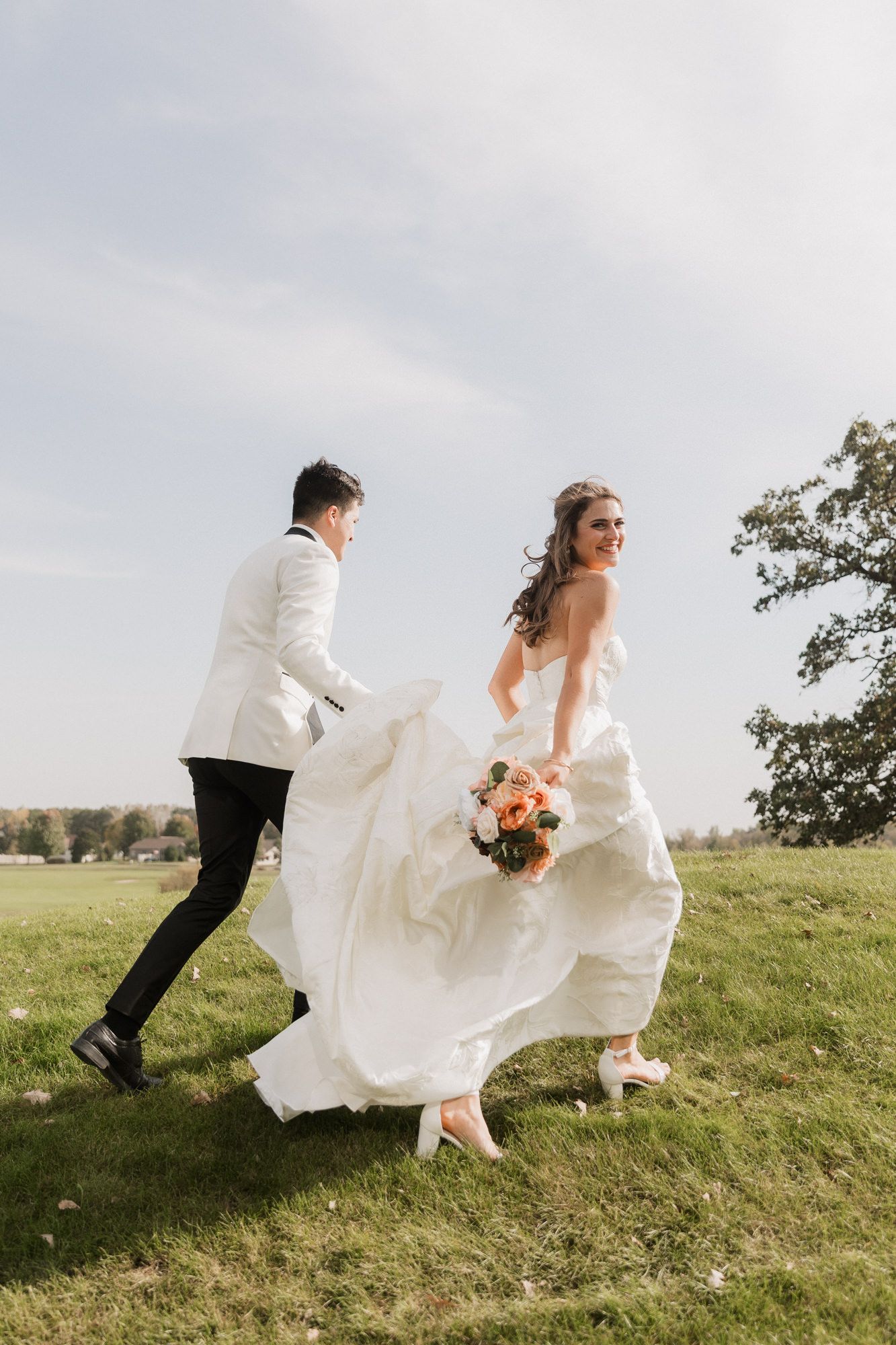 Bride and groom running joyfully across green field, bride holding floral bouquet in flowing white gown, groom in white jacket.