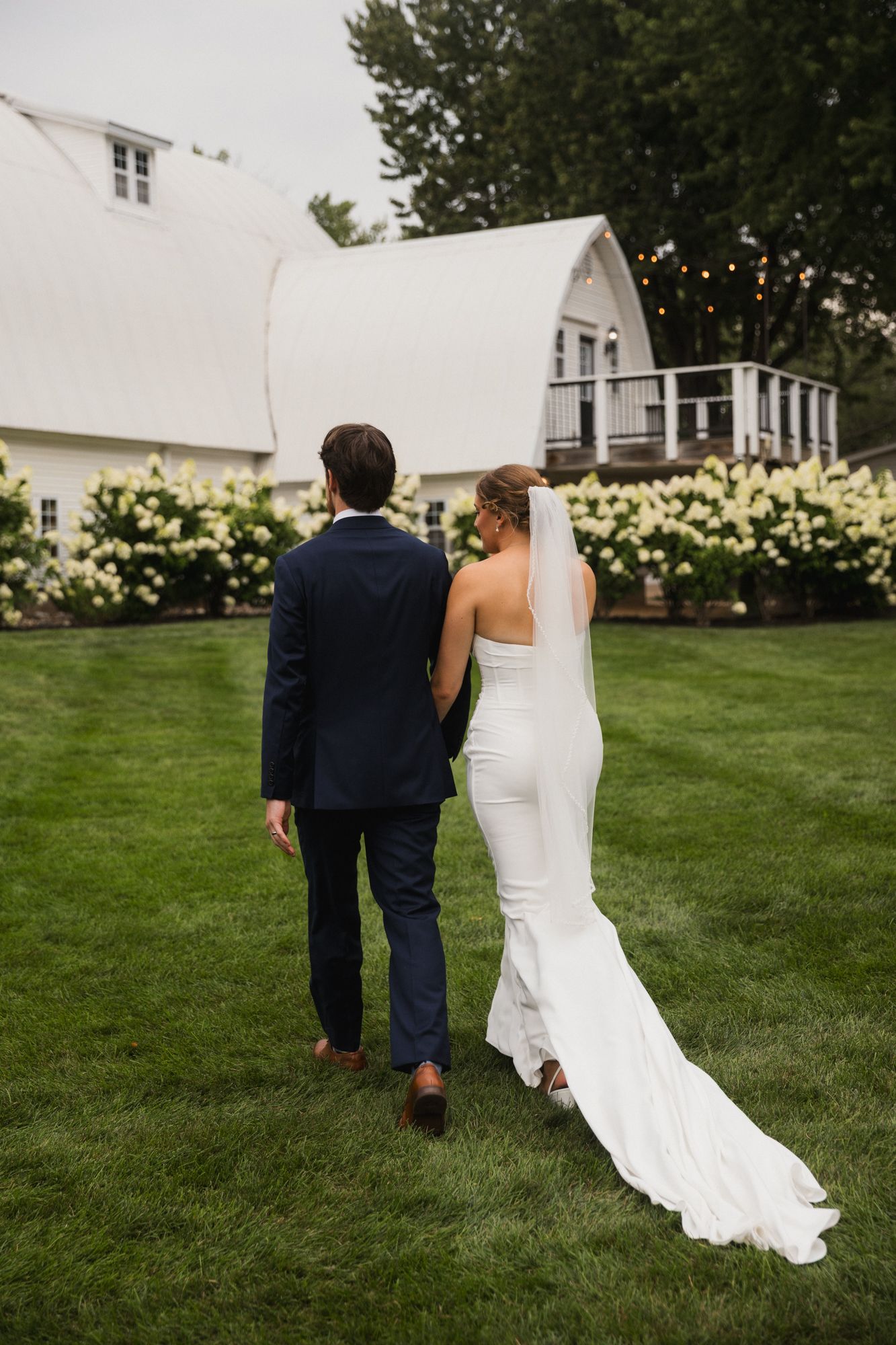 Bride and groom walking hand in hand across green lawn toward white barn venue surrounded by hydrangeas.