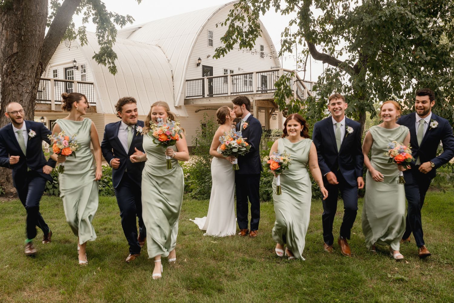 Wedding party walks outdoors in sage green bridesmaid dresses and navy suits, with bride and groom kissing in the center.