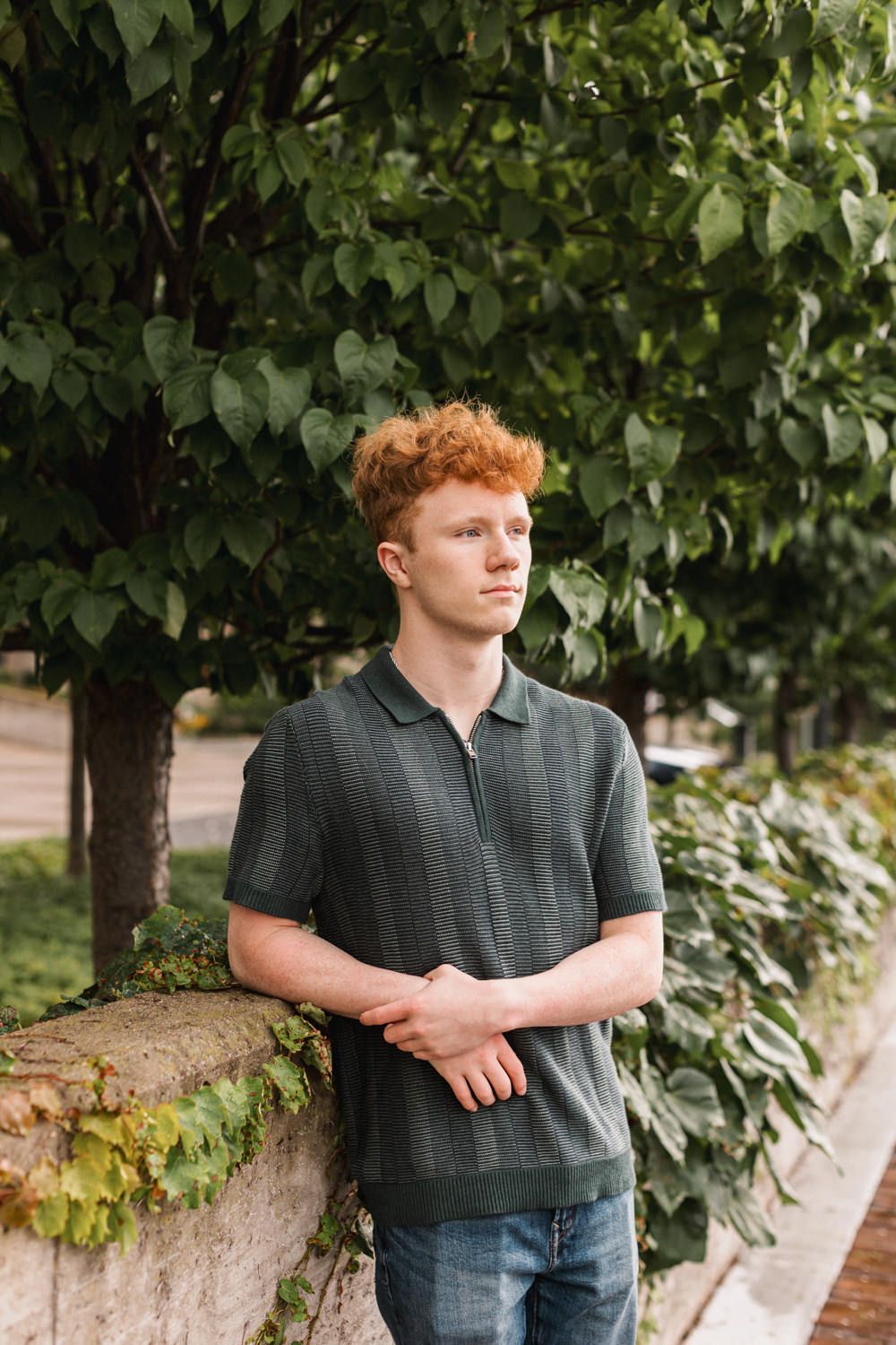 A person in a gray polo shirt poses against a leafy green backdrop in a casual outdoor setting.