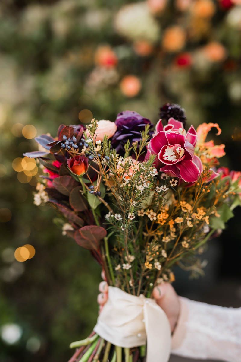 A row of colorful wedding bouquets held by hands with white sleeves against a blurred bokeh background.
