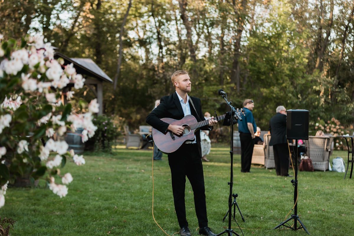 A wedding ceremony takes place outdoors in a garden setting with guests seated on white chairs at sunset.