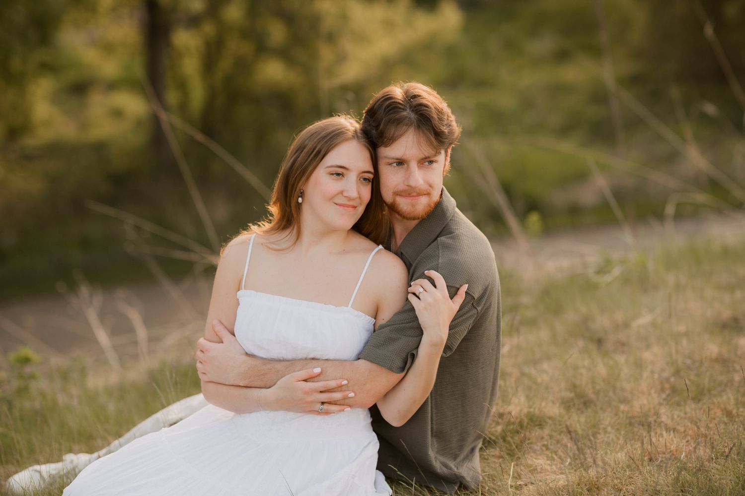 Couple embracing outdoors in golden hour light, woman in white dress, man in olive shirt, sitting in grassy field.