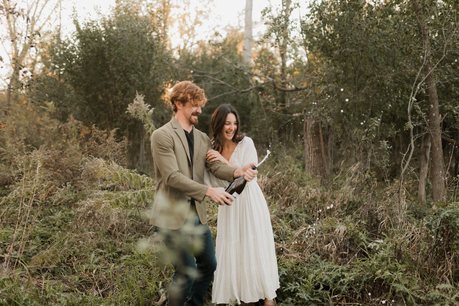 Couple smiling together in a lush forest setting, man in grey suit, woman in white dress.