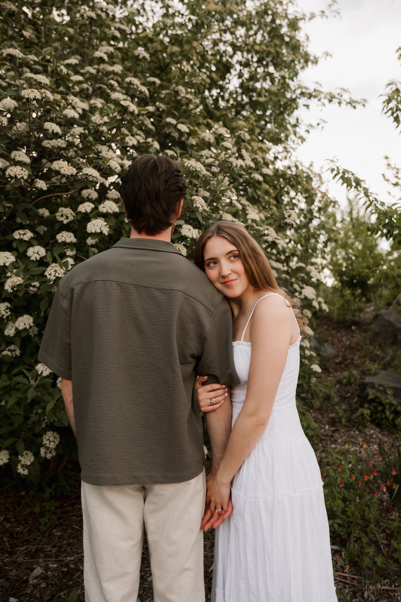 Woman in white dress holds mans arm near flowering bushes, looking back at camera.