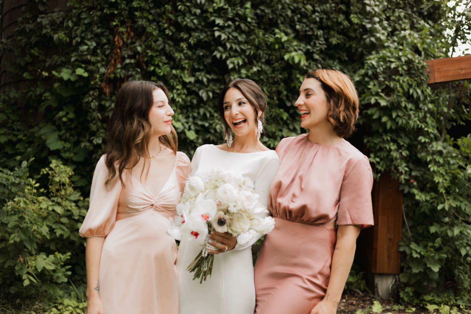 Wedding party members in soft pink dresses gather for photos in a garden setting.