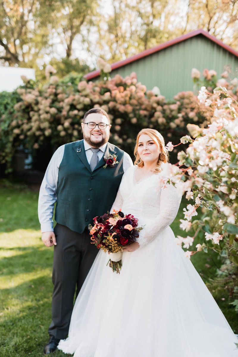 Newlyweds pose together in a garden setting with blooming trees and a rustic green building in the background.