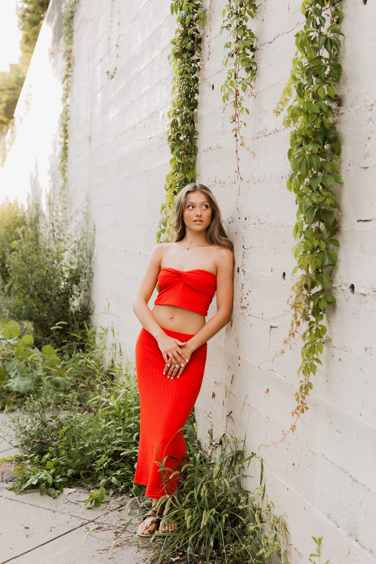 Young woman in red crop top and maxi skirt leaning against white brick wall with ivy, outdoor fashion portrait.
