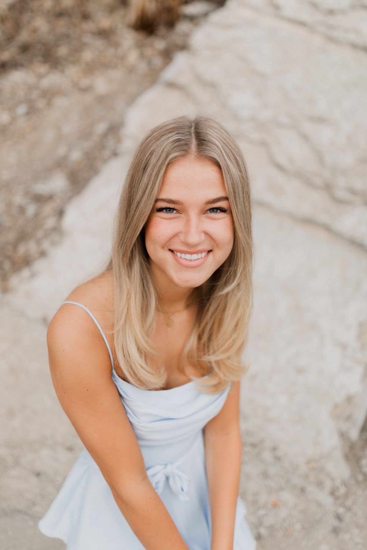 Smiling young blonde woman in a light blue dress posing outdoors against a rocky background.
