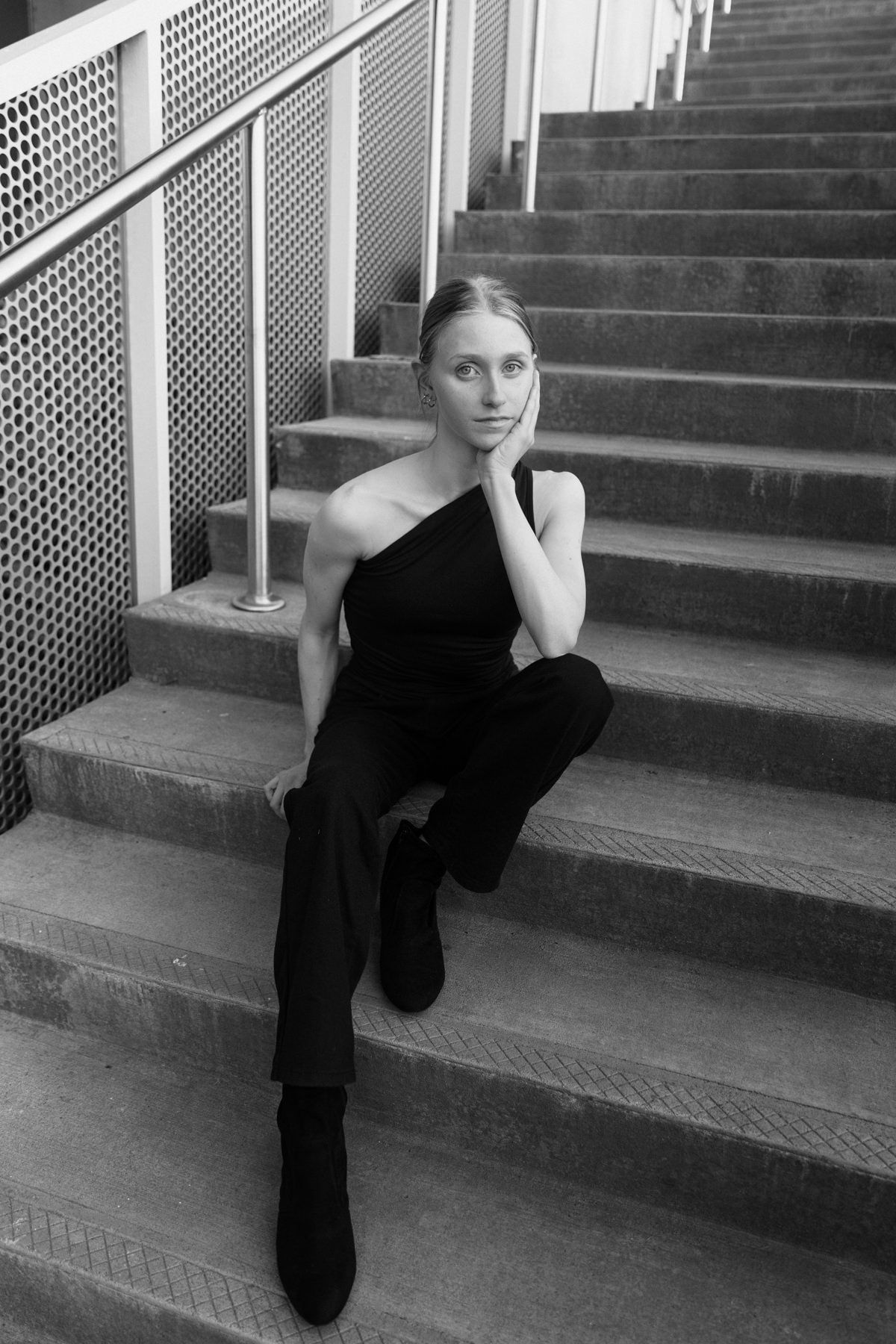 Woman in black one-shoulder jumpsuit sitting gracefully on outdoor stairs, resting chin on hand, black and white photo.