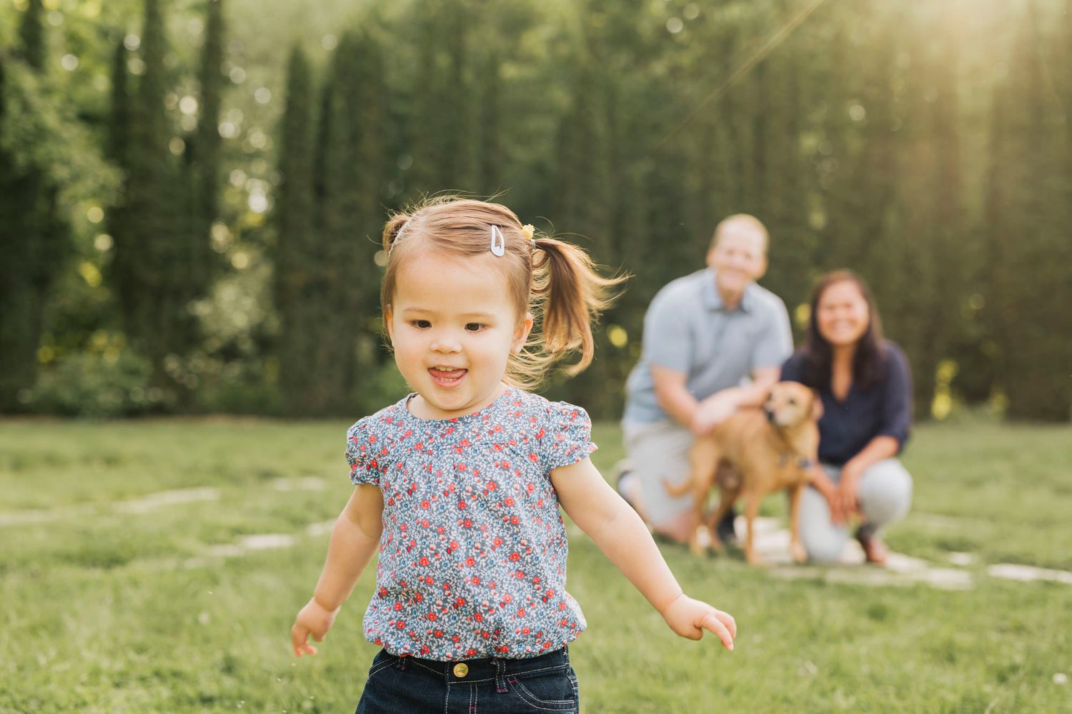 A toddler runs joyfully in a sunlit field while others sit in the background.