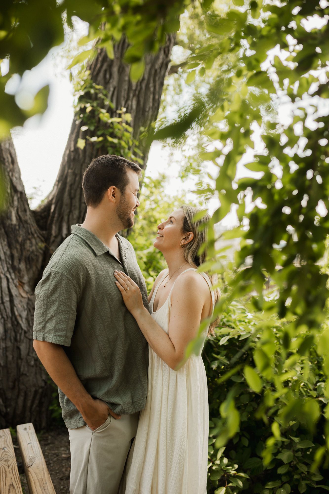 A couple shares a romantic moment under lush green trees, the woman in a white dress smiling up at the man.