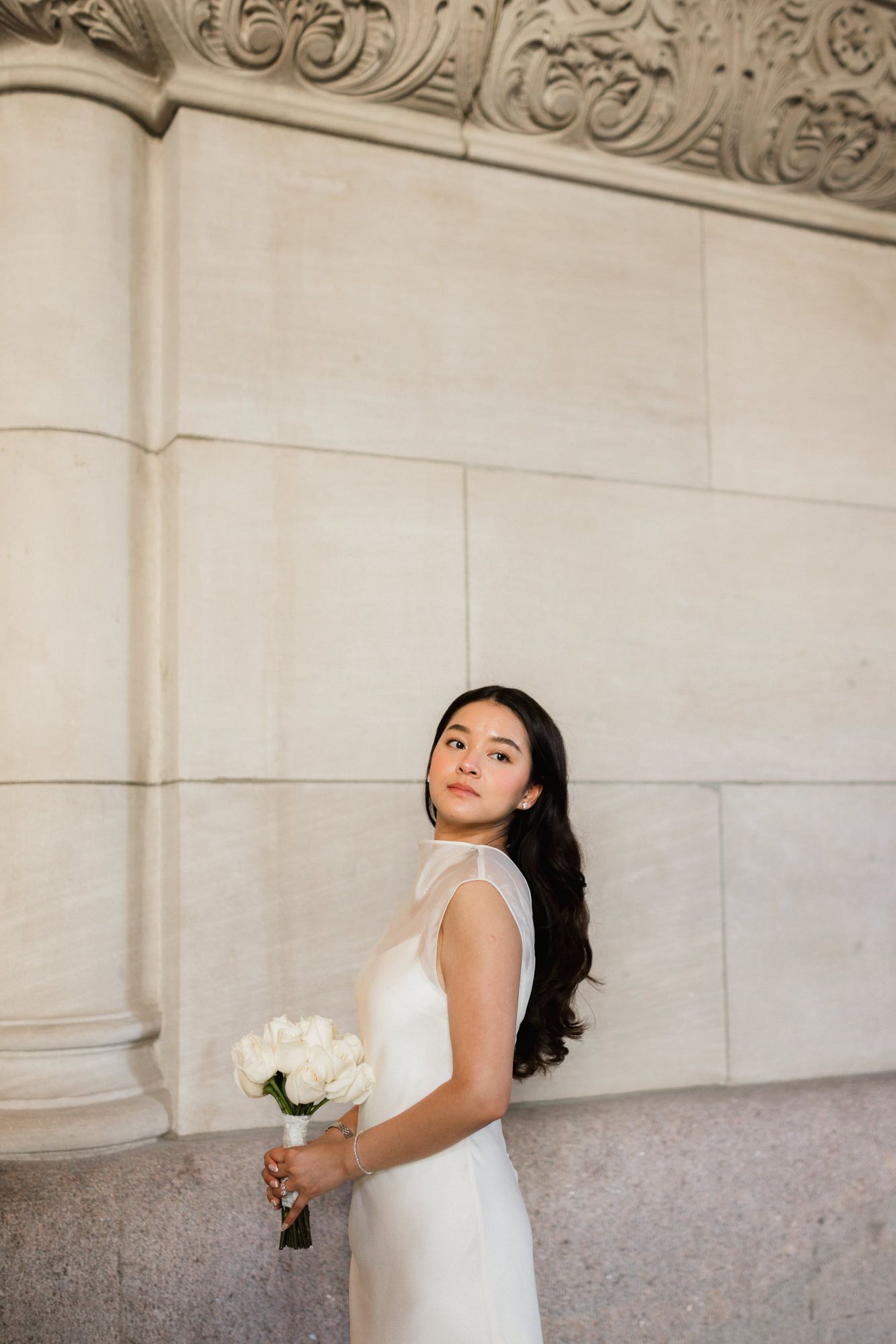 Elegant bride in white gown holding white roses bouquet, posing gracefully against ornate marble building exterior.