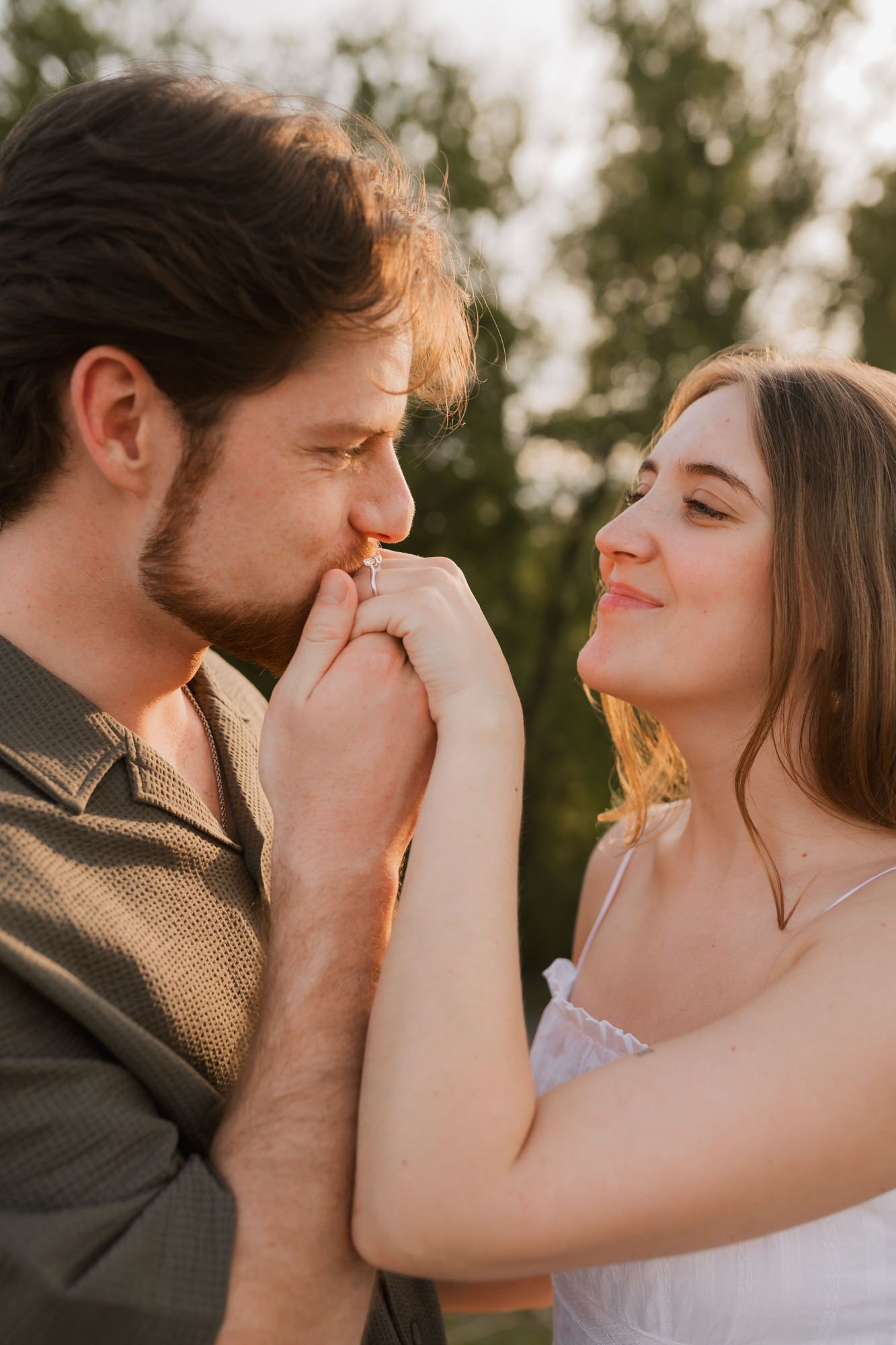 Man kisses womans hand as they face each other smiling in golden hour sunlight outdoors.