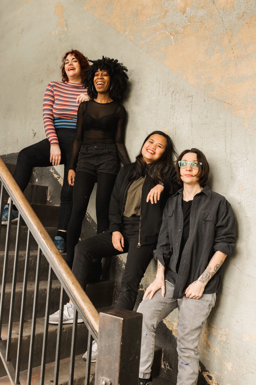 Four diverse young women laughing and posing on an urban staircase against a weathered wall.