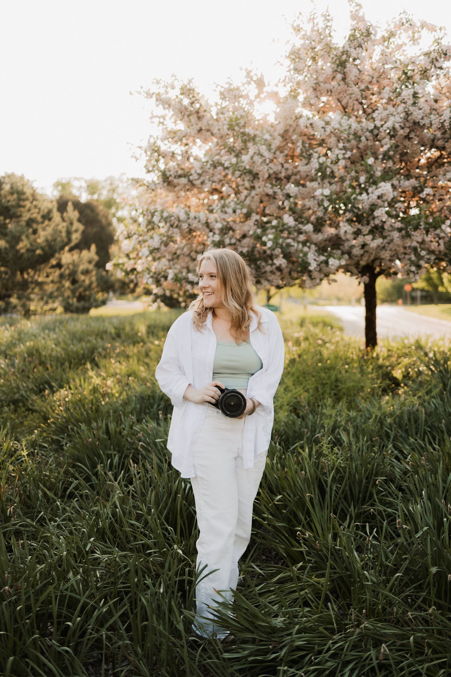 A blonde woman in white outfit holds a camera while standing in lush green grass near a blooming cherry tree.