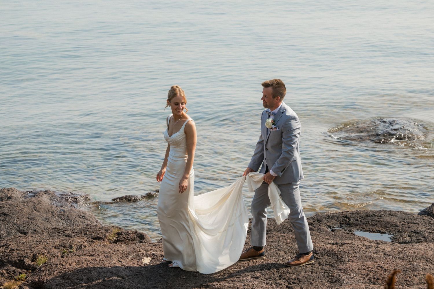 Bride and groom walk along rocky shoreline, groom carrying wedding dress train with ocean backdrop.