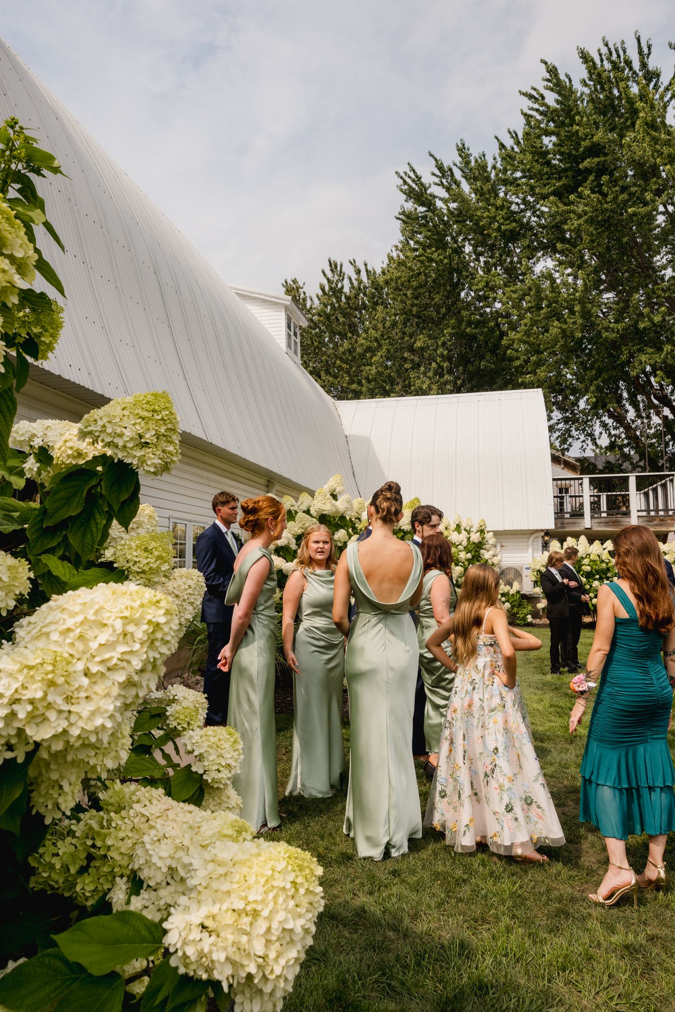 Bridesmaids in sage green dresses gather outdoors near white hydrangeas at an elegant barn wedding venue.