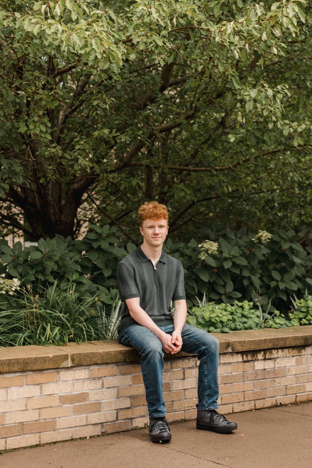 Person sitting on a brick wall outdoors with lush green trees and bushes in the background.