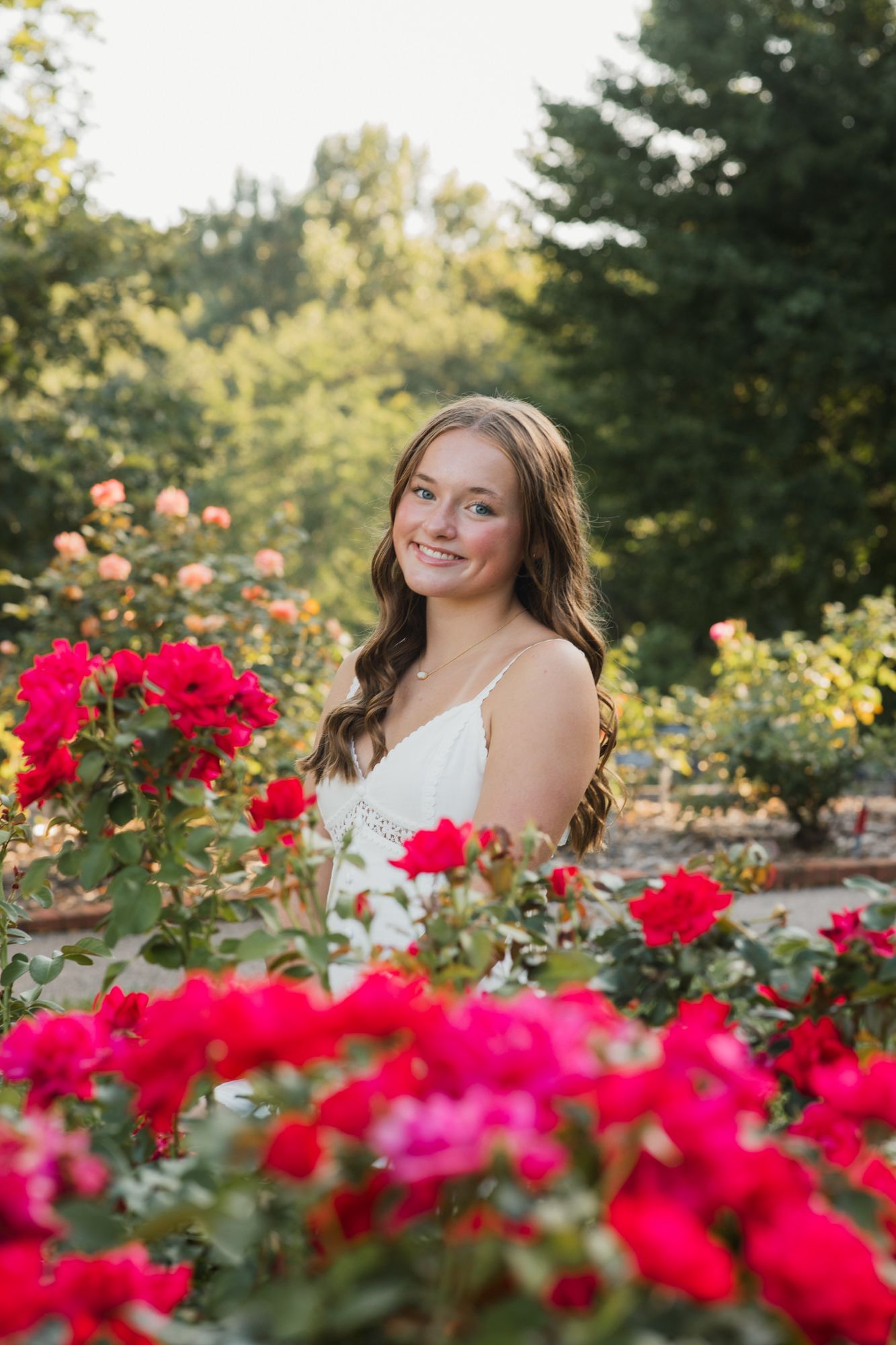 Young woman in white dress smiling among vibrant red and pink roses in a lush garden setting.