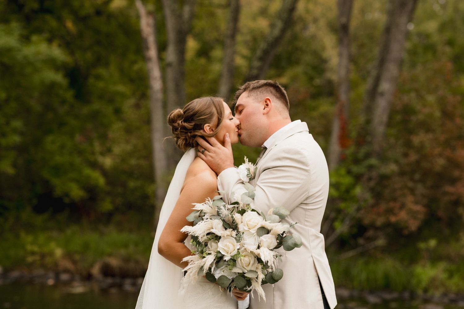 A bride and groom share a romantic kiss outdoors, surrounded by lush green trees, as she holds a white floral bouquet.