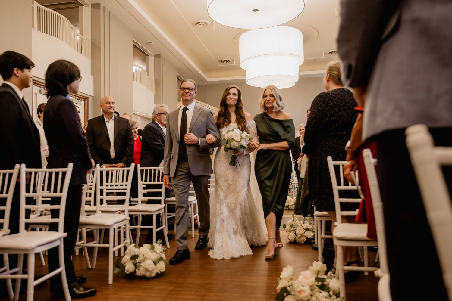 Bride walks down the aisle with parents at an elegant indoor wedding ceremony with white chairs and floral decor.