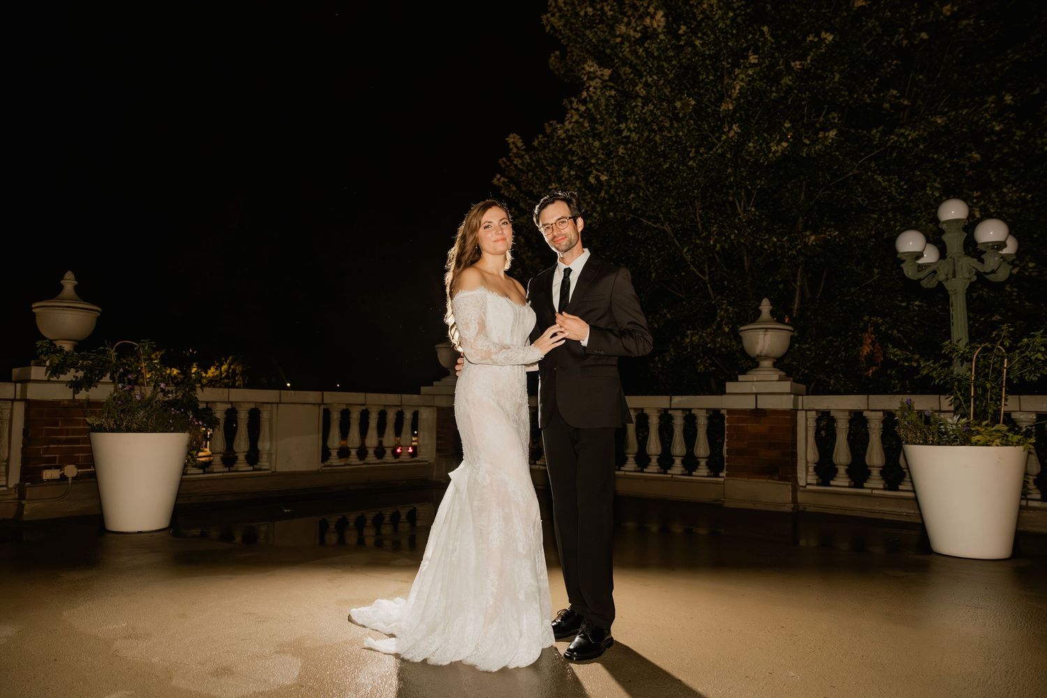Bride in white mermaid gown and groom in tuxedo share first dance on outdoor terrace at night.