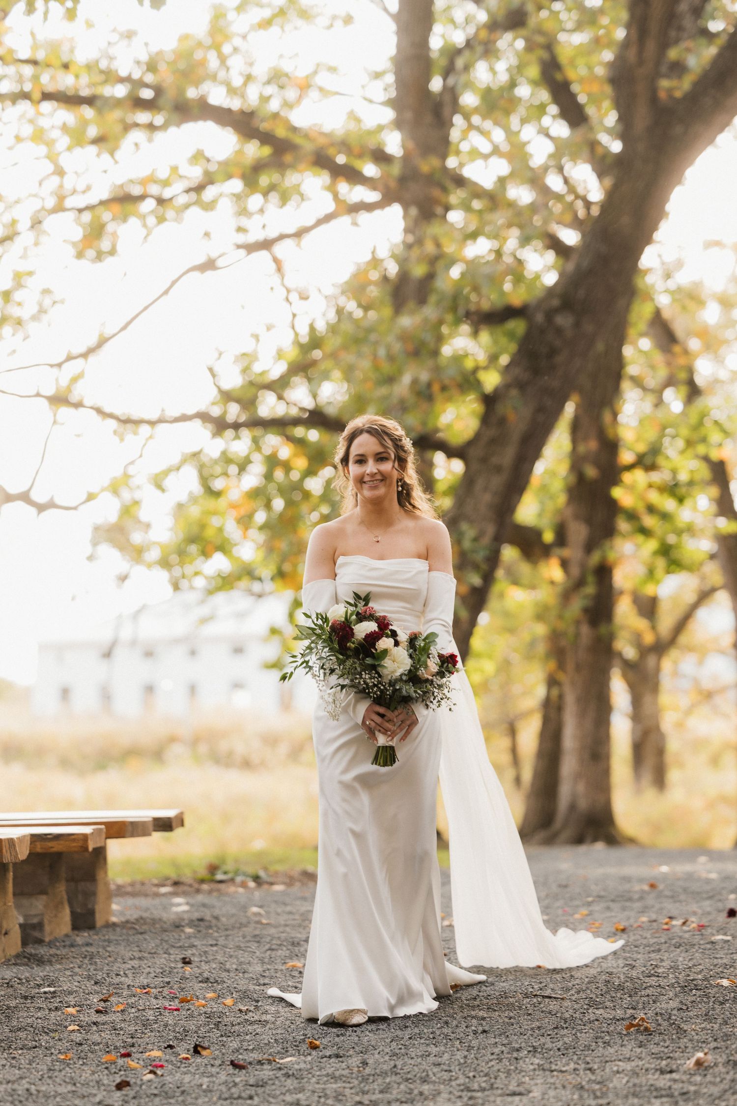 Bride in an off-shoulder satin wedding gown with sheer sleeves holds a burgundy and white bouquet while walking beneath autumn trees.