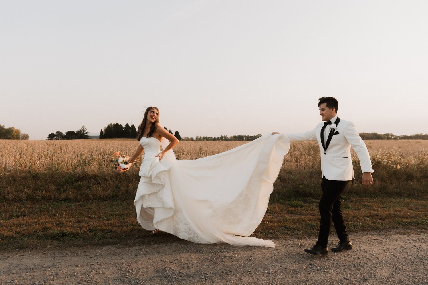 Bride and groom walk along a rural path at sunset, her white gown flowing as he holds the train.