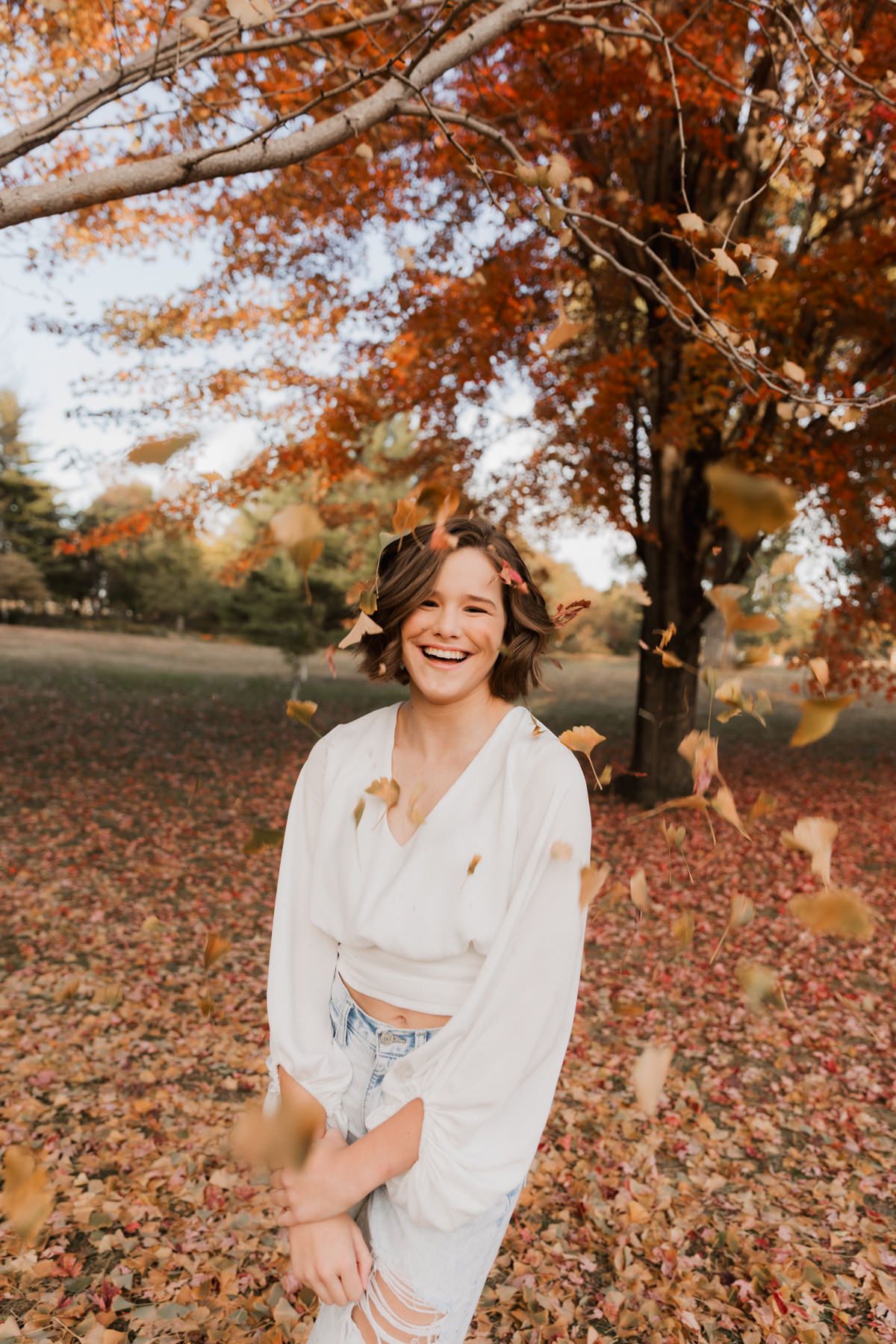 Laughing young woman in white outfit surrounded by falling autumn leaves in a colorful fall park.