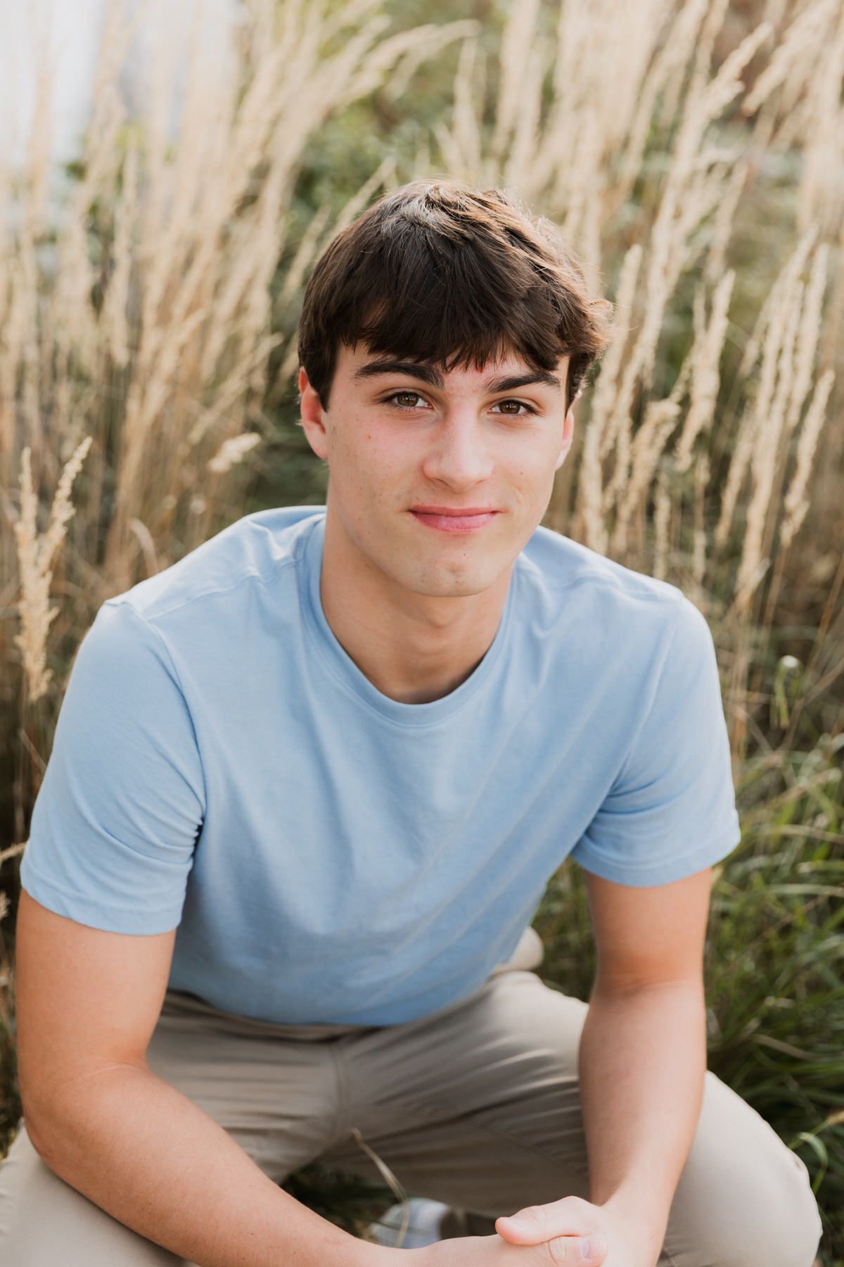Smiling teenage boy in light blue t-shirt sitting in front of tall golden grass outdoors.
