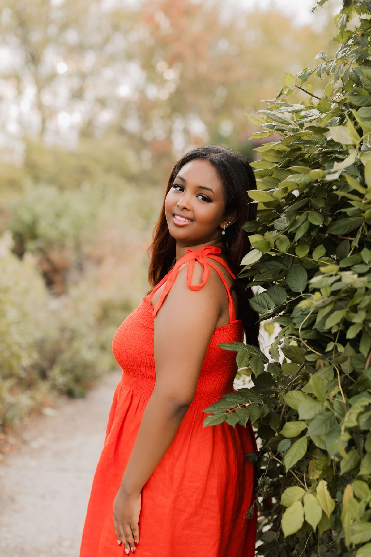 A smiling woman in a vibrant red dress poses beside lush green foliage on a garden path.