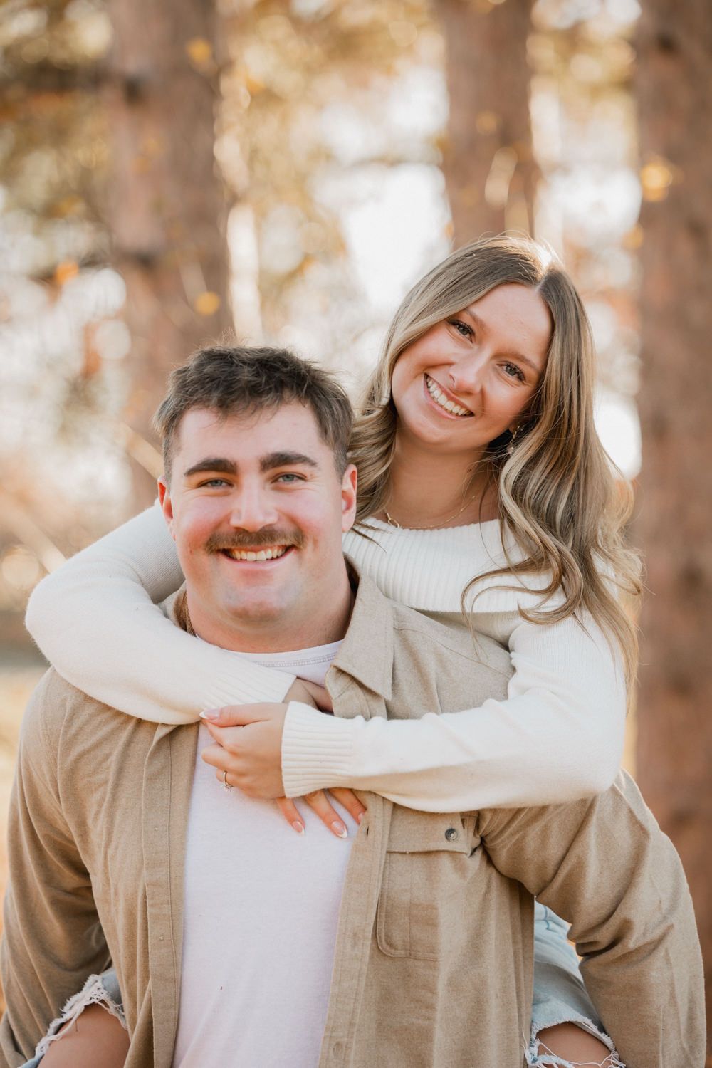 A couple embraces while sharing a piggyback ride in an autumn forest setting with warm golden light.