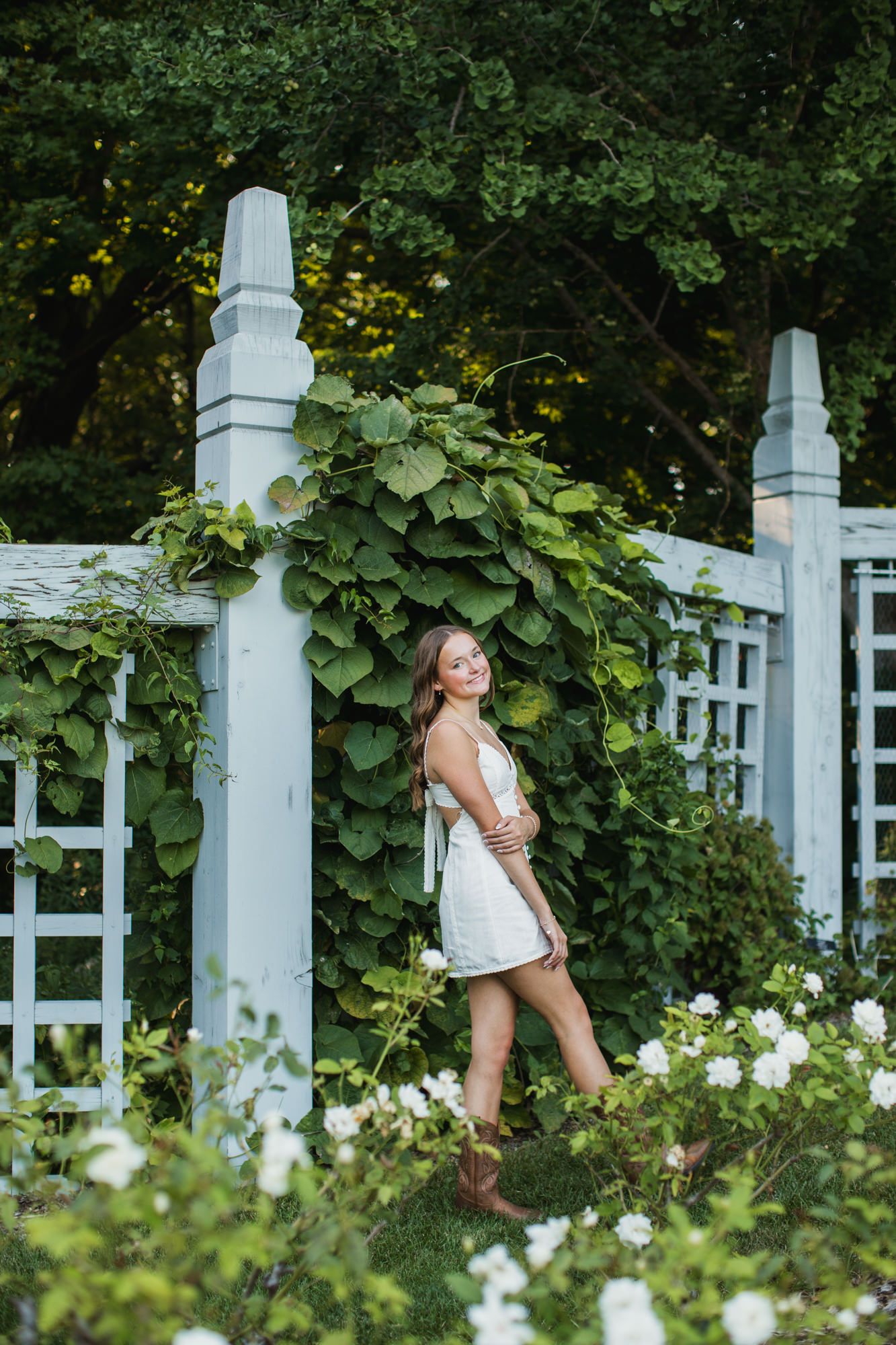 Young woman in white dress and boots poses by a vine-covered white garden trellis surrounded by white roses.