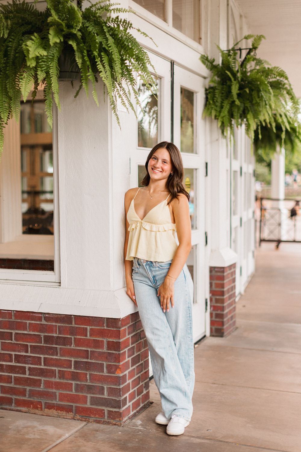 Woman in casual outfit poses on porch with hanging ferns and brick accents.