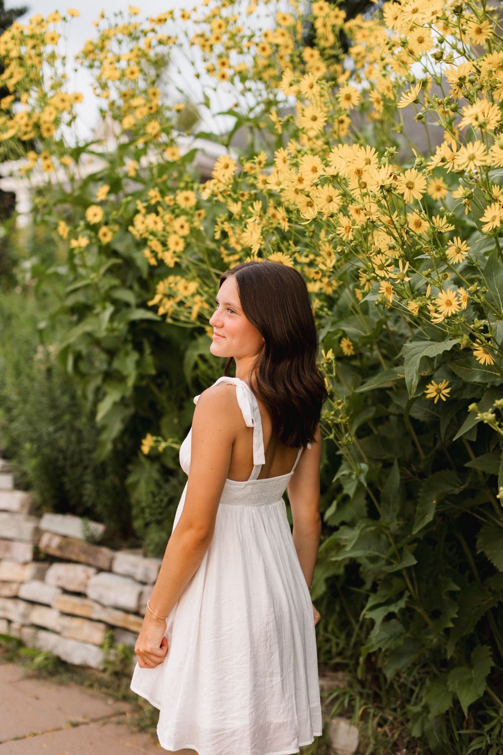 Woman in white sundress stands near blooming yellow wildflowers against stone wall on summer day.