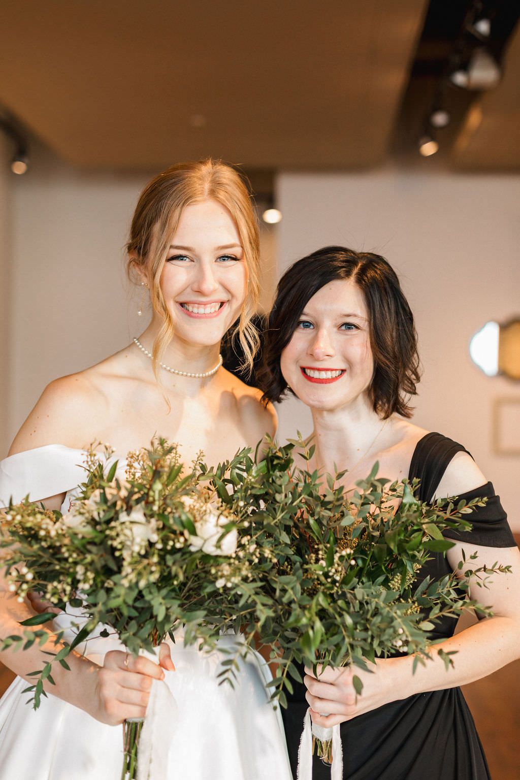 Bridesmaids in contrasting black and white dresses hold lush greenery bouquets while smiling at a wedding venue.