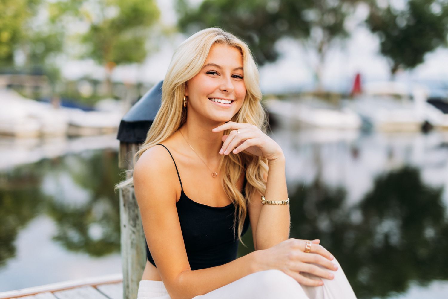 Person in black tank top smiling while sitting near marina with boats and reflective water in background.