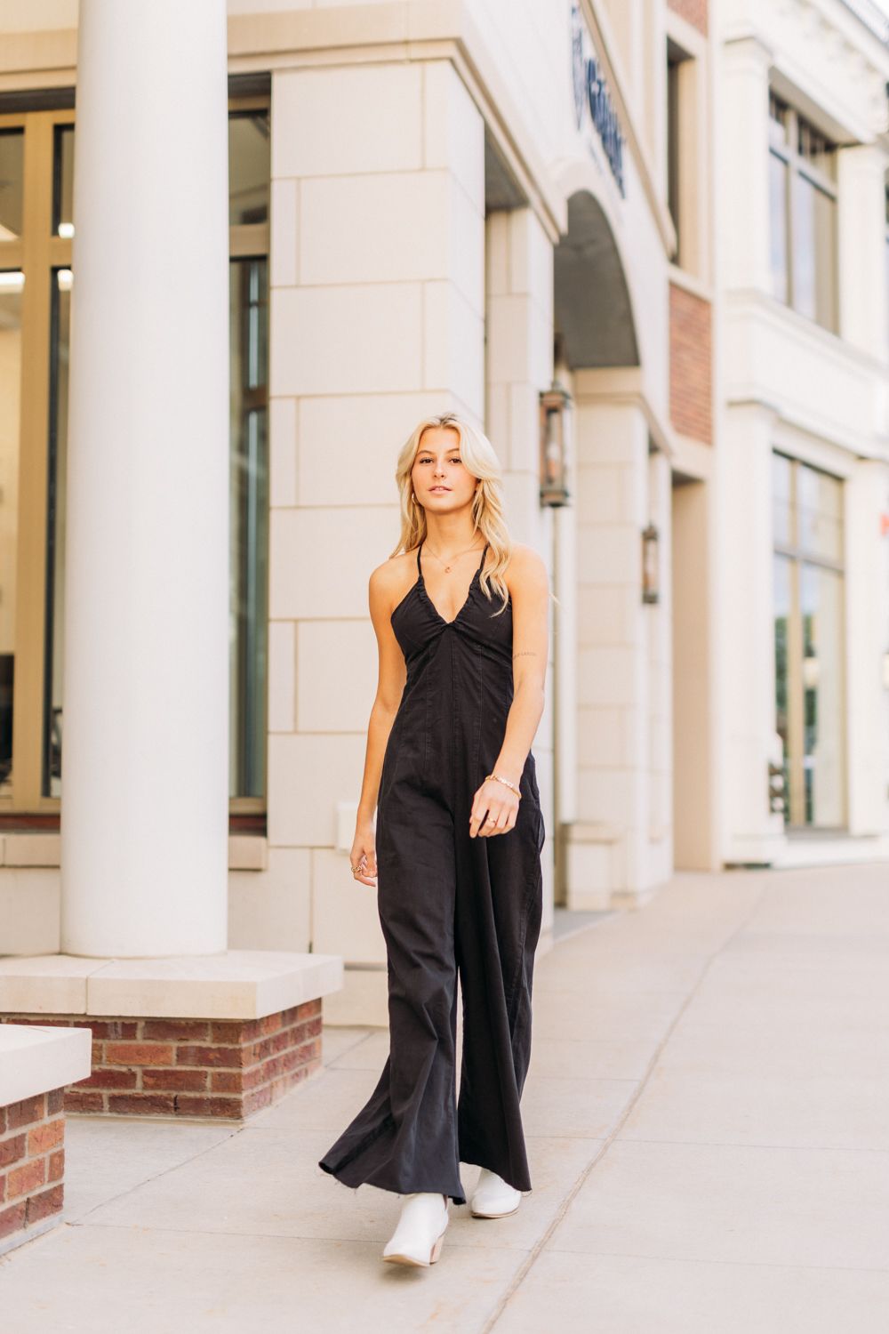 A person in a flowing black dress stands elegantly against white columns in an outdoor urban setting.