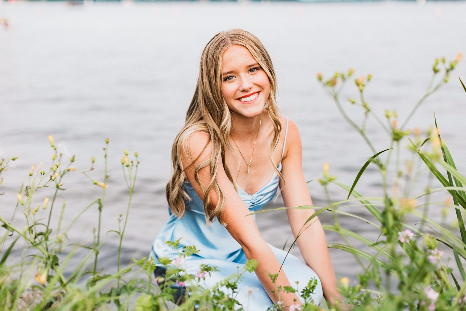 A person in a light blue dress crouches among wildflowers near the waterfront.