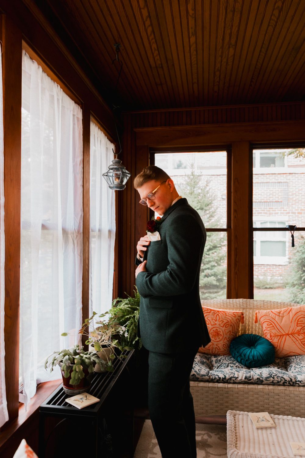 A groom adjusts his tie while standing in a sunlit enclosed porch with decorative pillows.