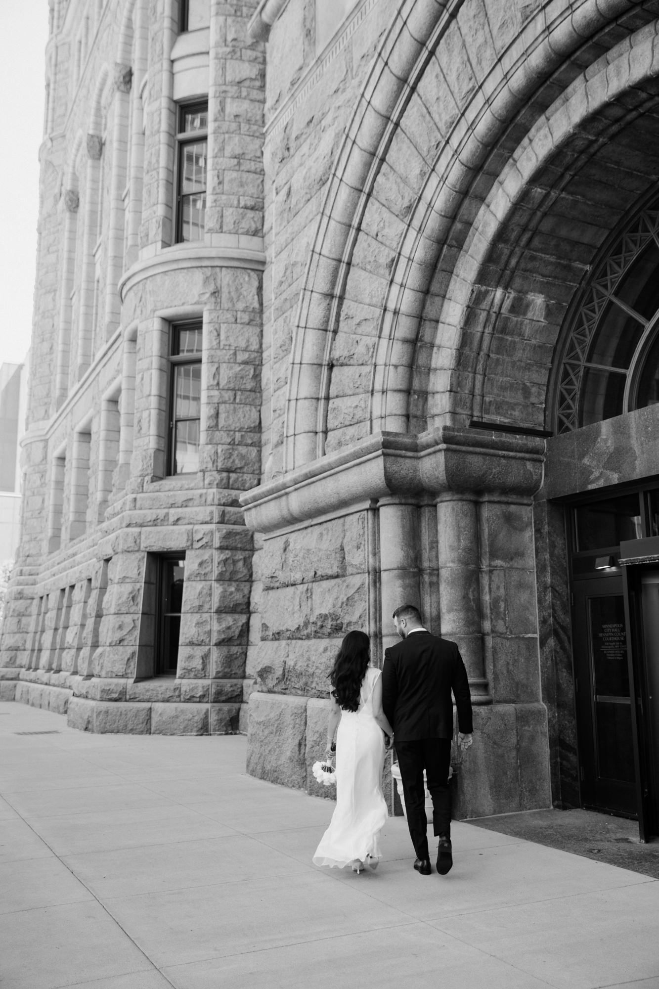 Black and white photo of a bride and groom walking toward a grand stone building entrance on their wedding day.