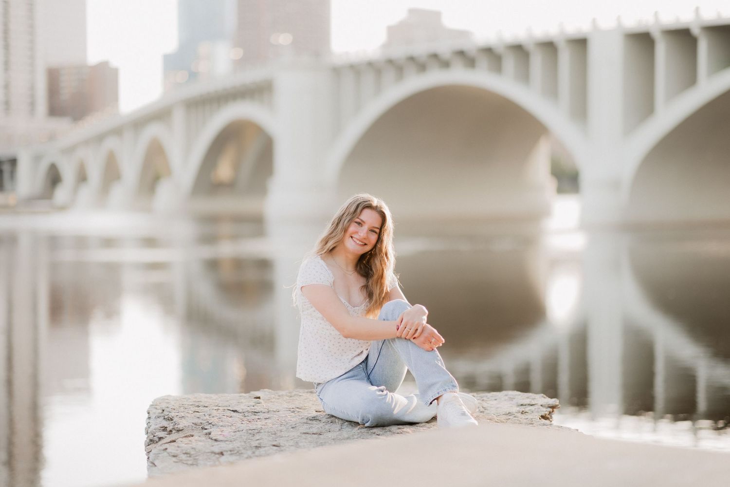 Young woman sitting on rocks by a river with an arched bridge reflected in calm water on a sunny day.