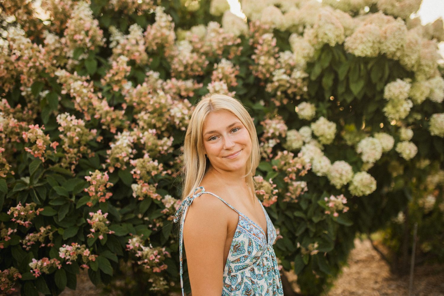 Smiling blonde woman in floral dress posing in front of blooming hydrangea bushes outdoors.