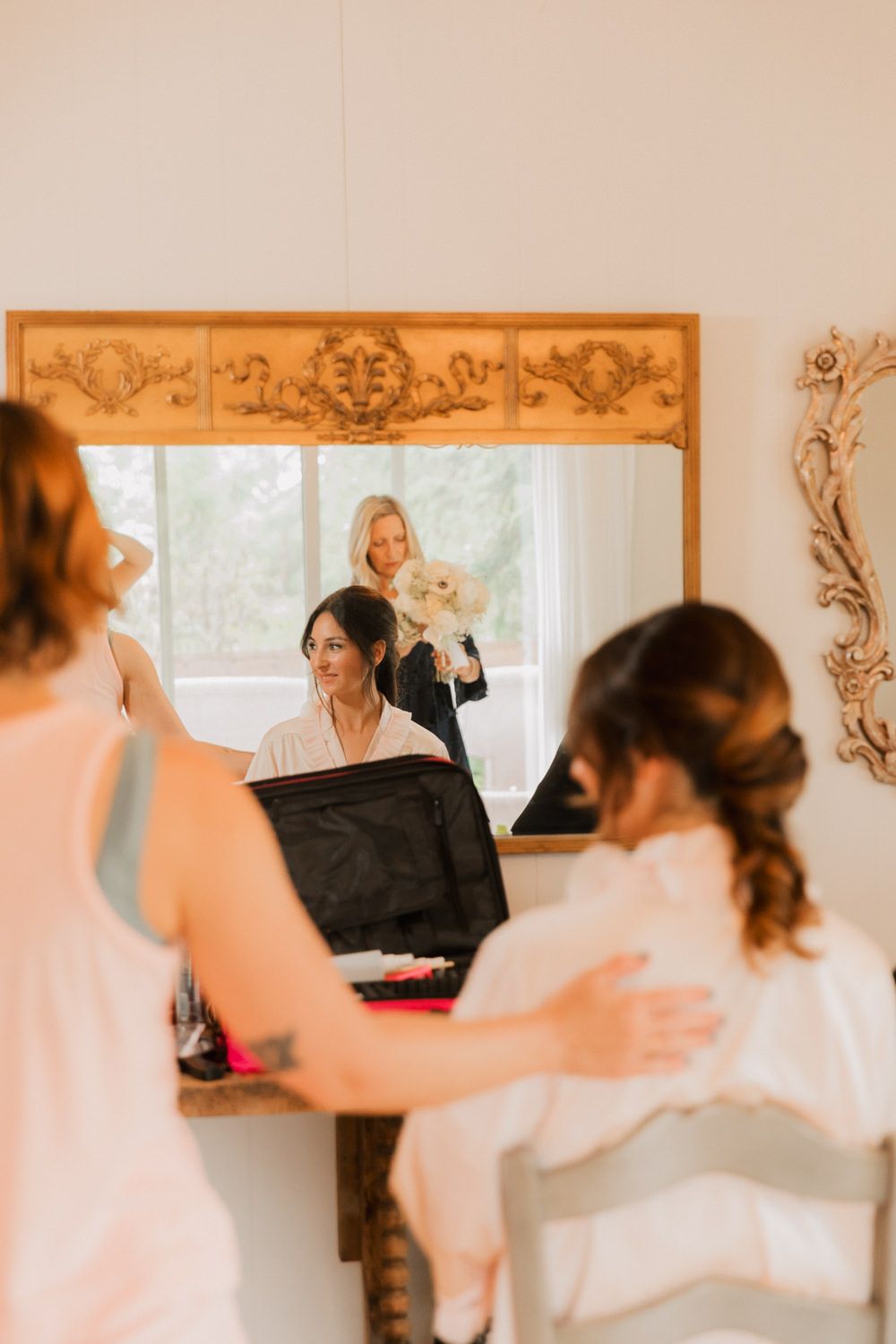 A reflection in an ornate mirror shows wedding preparations taking place in a sunlit room.