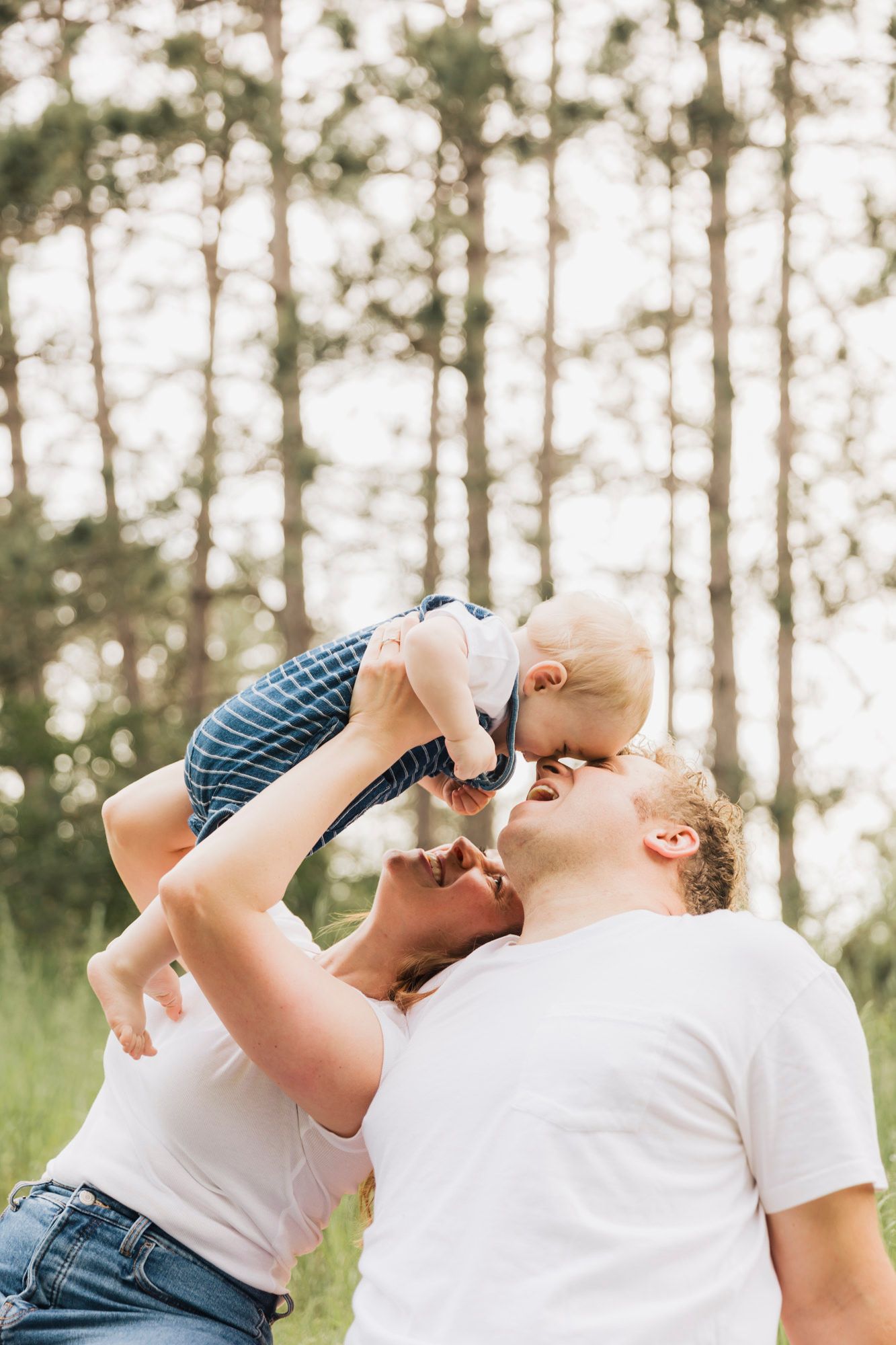 Parents playfully lift their baby in the air against a backdrop of tall pine trees.